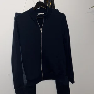 Svart zip hoodie med fickor - En stilren svart hoodie med hel dragkedja framtill och två fickor. Tröjan har en enkel design utan tryck, långärmad modell och ribbade muddar. Perfekt för lager-på-lager och chill dagar.