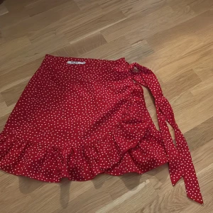 Röd prickig omlottkjol från NA-KD - Supersöt röd omlottkjol från NA-KD med vita prickar och volangkant. Kjolen knyts i sidan och har en feminin, lekfull vibe som passar perfekt till sneakers eller sandaler. Kort modell som ger en fräsch och somrig look.