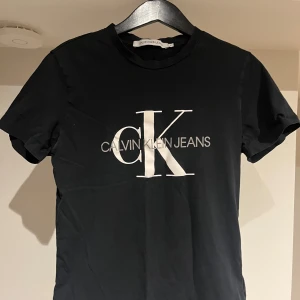 Svart t-shirt från Calvin Klein Jeans - Svart t-shirt från Calvin Klein Jeans med stor vit CK-logga och text på bröstet. Klassisk rund halsringning och korta ärmar. Skön passform och tillverkad i mjuk bomull, perfekt till jeans eller shorts. Bra skick! Bara att höra av sig vid eventuella frågor!😉