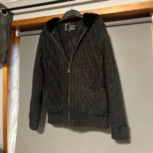 Grå stickad jacka med foder och päls - Snygg grå stickad jacka från Canda med quiltat foder och dragkedja. Jackan har en mysig huva med pälsdetaljer och ribbade muddar. Perfekt för kyliga dagar!