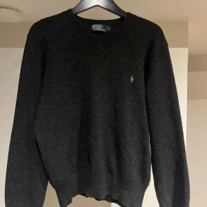 Svart ulltröja Polo Ralph Lauren - Mörkgrå, nästan svart, stickad tröja från Polo Ralph Lauren med klassisk v-ringning och broderad logga på bröstet. Tröjan är i mjuk ull och har långa ärmar samt ribbade muddar för en clean och tidlös look. Tröjan är i bra skick utan några defekter! Bara att höra av sig vid eventuella frågor!😉