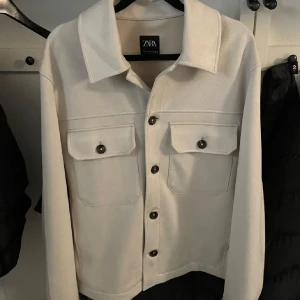 Jacka/ overshirt från Zara - Säljer en stilren vit overshirt från Zara i mycket bra skick. Jackan har en klassisk krage och knappar framtill. Två stora fickor med knappar på bröstet ger en snygg detalj. Perfekt för vår och höst men även inomhus på vintern. Använd 1 gång. Pris kan diskuteras.