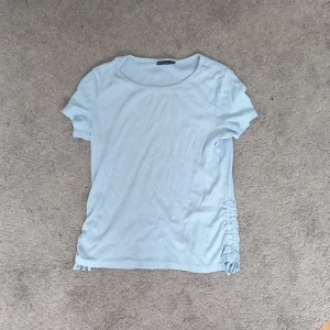 Ljusblå ribbad topp från XOXO Friends - Supersöt ljusblå ribbad topp från XOXO Friends med korta ärmar och snygg snörningsdetalj i sidan. Mjuk och stretchig, perfekt till jeans eller kjol. Enkel men ändå med en twist tack vare snörningen. Rund halsringning och figurnära passform.