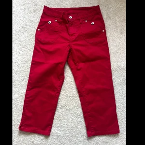 Röda capribyxor från Flash Jeans - Snygga röda capribyxor från Flash Jeans, modell Molly Capri. Byxorna har vita knappar, flera fickor fram och bak samt är tillverkade i stretchig bomull med lite elastan för skön passform. Perfekta för dig som vill sticka ut med färg.
