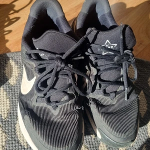 Svarta Nike sneakers med vit swoosh - Svarta Nike sneakers med vit swoosh och snörning. Ovandelen har ett ribbat mönster och är tillverkad i syntetmaterial. Skorna har en platt sula och rund tå, perfekt för dig som gillar sportig stil. Modellnummer DX7615-001.