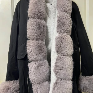 Svart parkajacka med fluffig fuskpäls - Parkajacka i svart med breda, ljusgrå fluffiga fuskpälsdetaljer längs krage, ärmslut och insida. Jackan har en oversized känsla och är perfekt för kalla dagar. Pälsen är supermjuk och ger jackan en unik look.