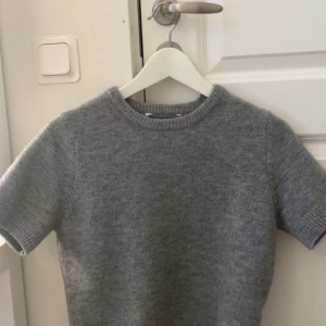 Grå stickad kortärmad tröja - Mysig grå stickad tröja med korta ärmar och rund hals. Perfekt för lager-på-lager och passar till både jeans och kjol. Enkel och stilren design med ribbade kanter vid hals, ärmslut och nederkant.
