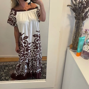 Brunvit offshoulder långklänning - Säljer en superfin långklänning i vitt och brunt med stora blommiga mönster. Klänningen har offshoulder-modell med resår upptill och korta puffärmar. Den är luftig och har volang nertill, perfekt för sommarens varma dagar.Helt ny,bara tvättad 