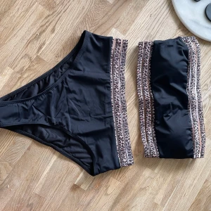 Svart bikini med leoparddetaljer - Snygg svart bikini med bandeau-topp och matchande trosa. Båda delarna har coola leopardmönstrade band i brunt och beige längs kanterna. Perfekt för stranden eller poolen om du vill sticka ut lite extra. Materialet är mjukt och stretchigt syntet för bästa komfort.Helt ny