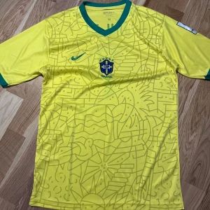 Brasilien Vini Jr. 7 Copa America tröja - Gul Brasilien fotbollströja från Nike med gröna detaljer, Vini Jr. och nummer 7 på ryggen. Copa America 2024-märke på ärmen. Unikt grafiskt mönster framtill och klassisk v-ringad krage. Tillverkad i lätt och ventilerande polyester.