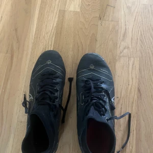 Svarta Nike fotbollsskor med guldiga detaljer elite  - Snygga svarta Nike fotbollsskor med guldiga detaljer och klassisk swoosh på sidan. Skorna har snörning och mönstrad ovandel för extra grepp. Perfekta för dig som vill sticka ut på planen med stilrena och sportiga skor.