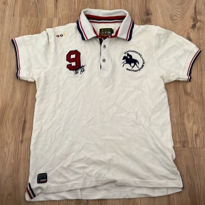 Vit pikétröja från Huntington Polo Club - Vit pikétröja från Huntington Polo Club med broderad logga och stor röd siffra 9 på bröstet. Krage och ärmslut har snygga ränder i rött, vitt och blått. Klassisk piké med korta ärmar och sportig känsla, perfekt till jeans eller shorts.
