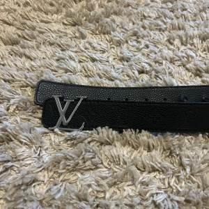 Svart Louis Vuitton skärp i skinn - Snyggt svart skärp från Louis Vuitton i äkta skinn med klassisk LV-logga i silvermetall framtill. Bältet har präglat monogrammönster längs hela remmen och en stilren, rektangulär form. Perfekt accessoar för att lyfta din outfit. 105 cm i längden 