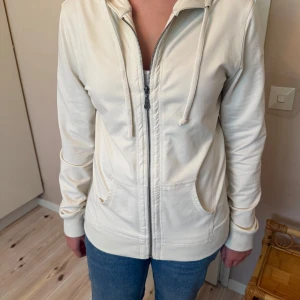 Vit hoodie från Timberland med dragkedja - Vit hoodie från Timberland i bomull med broderad logga på bröstet. Tröjan har huva med snörning, två fickor fram och hel dragkedja. Långärmad och med ribbade muddar för en skön passform. Perfekt för chill dagar eller när du vill ha en clean look.