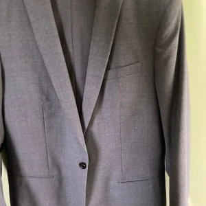 Grå kavaj från Filippa K, storlek 54 - Stilren grå kavaj från Filippa K i klassisk modell med smal krage och enkelknäppning. Kavajen har tre fickor framtill och är fodrad med slät insida. Endast kavaj. 