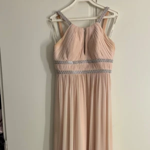 Rosa långklänning med paljetter  - Elegant långklänning i rosa färg från By Kris. Klänningen har smala axelband med glittriga detaljer, plisserad överdel och markerad midja med strassband. Flödande kjol i tunt, svepande material som ger en lyxig känsla. Perfekt för bal eller fest. Klänningen är lite fläckig, som visas på sista bilden men som inte syns vid användning:)