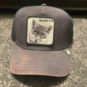 Svart truckerkeps Silver Fox The Farm - Cool svart truckerkeps från The Farm med broderad patch framtill med en silverräv och texten 'SILVER FOX'. Kepsen har mesh på sidorna och bak för extra ventilation och böjd skärm. Perfekt för dig som gillar djurmotiv och streetstyle.