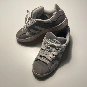 Grå Adidas Campus sneakers - Snygga adidas Campus sneakers i grå mocka med vita detaljer och klassiska tre ränder på sidorna. Skorna har vit sula med gummiyta undertill och Ortholite-innersula för extra komfort. Perfekt för dig som gillar retrostil och streetwear. Något skavd inuti på ena, se bild 