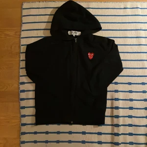 Svart hoodie Comme des Garçons Play S - Svart hoodie från Comme des Garçons Play i storlek S. Tröjan har dragkedja, huva och det ikoniska röda hjärta-emblemet med ögon broderat på bröstet. Perfekt för dig som gillar streetwear och vill sticka ut med en stilren look.