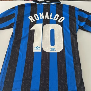 Inter Ronaldo 10 Umbro fotbollströja - Klassisk Inter fotbollströja med Ronaldo 10 på ryggen. Tröjan är blå och svart med vertikala ränder, broderat Inter-märke och Pirelli som sponsor på bröstet. Tillverkad i lätt och ventilerande polyester, med vit Umbro-logga och krage med svarta och vita detaljer. Endast handtvätt gäller. Replika
