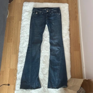 True Religion lowrise bootcut jeans - Säljer ett par snygga blå jeans från True Religion med bootcut passform och låg midja. Har klassiska kontrastsömmar, stora bakfickor med lock och vita knappar. Tillverkade i bomull med en skön stretch.