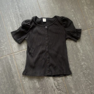 Svart ribbad blus från Lindex - Svart ribbad blus från Lindex med kort puffärm och rund halsringning. Toppen har knappar framtill och är tillverkad i ett mjukt, stretchigt material som sitter snyggt. 