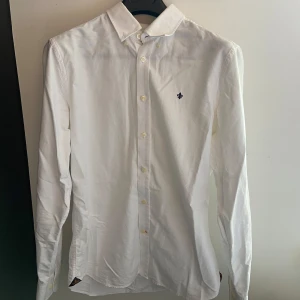 Vit skjorta från Morris Stockholm M - Klassisk vit skjorta från Morris Stockholm i storlek M. Skjortan har lång ärm, knappar framtill och en liten broderad logga på bröstet. Tillverkad i bomull med twillstruktur, perfekt för en clean och stilren look.