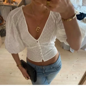 Vit broderad blus med puffärm - Superfin vit blus med broderade hålmönster, croppad passform och djup v-ringning. Puffiga trekvartsärmar och knappar framtill ger en romantisk vibe. Perfekt till jeans för en trendig look.
