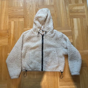 Beige teddyjacka med huva från H&M - Supermjuk och fluffig croppad teddyjacka från H&M Divided i beige. Jackan har huva, hel dragkedja framtill och snörning i nederkant för en snygg passform. Perfekt för kyliga dagar och riktigt cozy vibes.