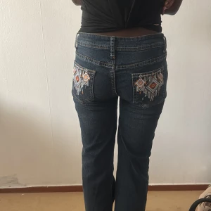 Mörkblå jeans med broderade bakfickor - Snygga mörkblå jeans med raka ben och coola broderade detaljer i orange och vitt på bakfickorna. Klassisk femficksmodell med kontrastsömmar och normal passform. Perfekta för dig som vill sticka ut lite extra.