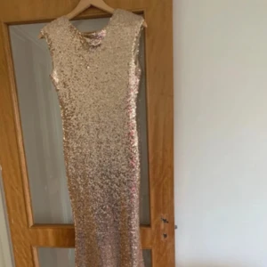 Glittrig långklänning i champagne - Säljer en maxad långklänning i champagnefärg med glittriga paljetter över hela klänningen. Den har ärmlös design, rund halsringning fram och djup v-ringning i ryggen. Perfekt för dig som vill sticka ut på festen!