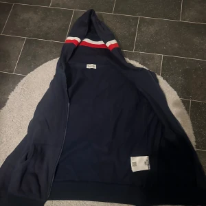 Blå Moncler hoodie med dragkedja - Snygg marinblå hoodie från Moncler med dragkedja och huva. Ärmarna har coola ränder i rött och vitt vid mudden och på insidan av huvan. Klassisk Moncler-logga på ärmen. Perfekt för dig som gillar streetwear och vill ha något stilrent men ändå sportigt.