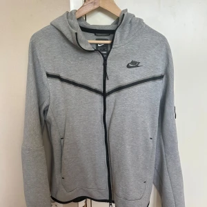 Nike tech hoodie - Snygg grå hoodie från Nike med hel dragkedja och svarta detaljer. Jackan har huva, långa ärmar och praktiska fickor med dragkedja. Perfekt för chill eller träning, med klassisk Nike-logga på bröstet och sportig look.
