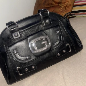 Svart handväska från Gesto med mönster - Cool svart handväska från Gesto i skinnimitation med präglat blommönster. Väskan har silvriga metalldetaljer, stor G-logga framtill och dubbla handtag. Praktisk dragkedja på baksidan och snygga nitar framtill. Perfekt för dig som gillar statement-accessoarer.