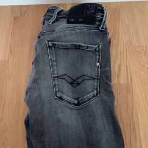 Replay Anbass grå jeans - Snygga Replay Anbass jeans i tvättad grå färg med klassisk femficksdesign och coola detaljer på bakfickan. Jeansen har en smal passform och är tillverkade i stretchigt denim för extra komfort. Perfekta för dig som gillar en modern och avslappnad stil.