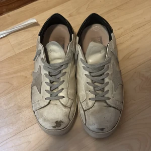 Grå/vita sneakers med stjärndetalj - Snygga sneakers från Golden Goose i vitt och grått skinn med ikonisk stjärna på sidan. Grå snörning och svart detalj vid hälen. Klassisk platt sula och rund tå, perfekt för en avslappnad streetstyle-look. Storlek 45 och skick 7/10. Har också två små skador under båda skorna men det går att fixa enkelt. Pris 1000 men kan pris förhandla lite.