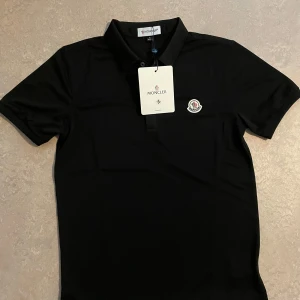 Svart piké Moncler - Snygg svart piké Moncler. Tröjan har Monclers ikoniska logga broderad på bröstet och stängs med knappar framtill. Tillverkad i mjuk bomull som känns skön mot huden.                                    OBS. Den är ny utan prislapp. Och liten i storleken faktiskt.