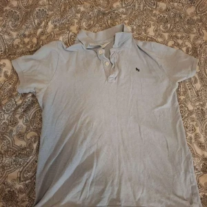 Ljusblå pikétröja med krage - Ljusblå pikétröja med klassisk krage och tre knappar framtill. T-shirten har korta ärmar och en liten broderad logga på bröstet. Perfekt för en chill och stilren look.