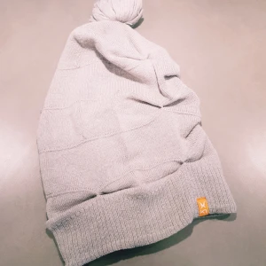 Ljusgrå mössa med tofs från Kari Traa - Ljusgrå stickad mössa från Kari Traa med ribbad kant och en stor tofs på toppen. Klassisk och enkel design med diskret orange logga på kanten. Perfekt för kalla dagar och snygg till vinterjackan.  Lite tråddragningar annars helt & fint skick