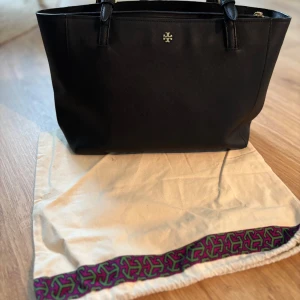 Svart handväska från Tory Burch - Stilren svart handväska från Tory Burch i skinn med klassisk rektangulär form och guldfärgad logga framtill. Väskan har två handtag, dragkedja upptill och rymligt foder med innerfack. Perfekt för dig som gillar en clean och tidlös look.