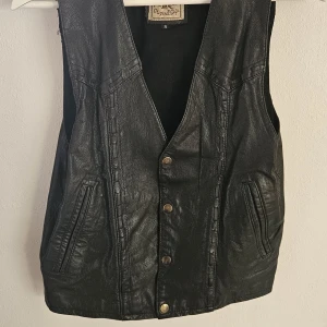 Svart skinnväst från Original Style - Svart skinnväst från Original Style med cool western-vibe. Västen har tryckknappar i mässing, två fickor framtill och markerade sömmar som ger extra edge. Perfekt att slänga över en hoodie eller skjorta för en grym look.