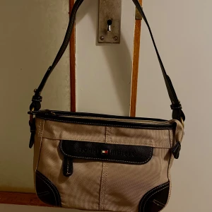 Beige axelväska Tommy Hilfiger - Snygg beige axelväska från Tommy Hilfiger med svarta detaljer och logga framtill. Väskan har justerbar axelrem, dragkedja upptill och svarta skinnpartier på kanter och fickor. Perfekt storlek för det viktigaste och stilren design som lyfter varje outfit. Inte fast pris, kan diskuteras 