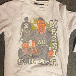 Vit Messi G.O.A.T. sweatshirt - Vit sweatshirt med stort tryck av Messi i PSG-tröja, texten 'MESSI' längs sidan och 'G.O.A.T.' längst ner. Tröjan har rund hals och långa ärmar. Perfekt för dig som älskar fotboll och vill hylla en av de största spelarna.