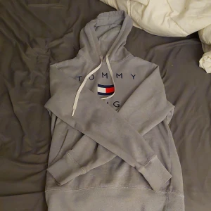 Grå hoodie Tommy Hilfiger XS - Säljer en grå hoodie från Tommy Hilfiger i storlek XS. Tröjan har klassisk huva med snörning och logga samt flagga broderad på bröstet. Mjuk och skön, perfekt för chill dagar. Ribbade muddar vid ärmslut och nederkant.