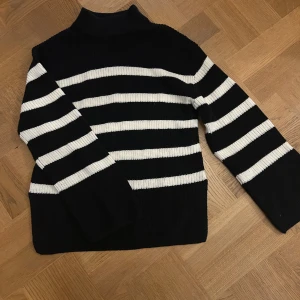 Randig svartvit stickad polotröja - Snygg stickad polotröja med breda svarta och vita ränder. Tröjan har hög krage och långa ärmar, perfekt för kyliga dagar. Den ribbade strukturen ger en skön och avslappnad känsla. Passar dig som gillar klassisk stil med en modern twist.