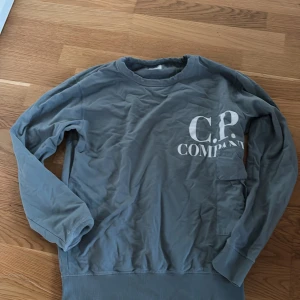 Grå sweatshirt från C.P. Company - Snygg grå sweatshirt från C.P. Company med stor vit logga på bröstet och en cool ficka på ena ärmen. Tröjan har rund hals och långa ärmar, perfekt för en avslappnad streetstyle-look. Tillverkad i mjuk bomull för skön känsla hela dagen.