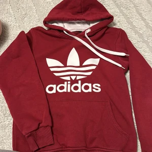 Röd Adidas hoodie med vit logga - Säljer en snygg röd hoodie från Adidas med stor vit Trefoil-logga och text på bröstet. Tröjan har huva med vita snören och en stor magficka. Perfekt för dig som gillar sportig och avslappnad stil.