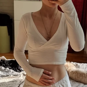 Vit omlott croppad långärmad topp - Säljer en vit croppad topp med omlott och djup v-ringning. Toppen har långa ärmar och är tillverkad i ett mjukt, stretchigt material som sitter snyggt på kroppen. Perfekt för dig som gillar en enkel men trendig stil.