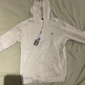 Grå hoodie från Polo Ralph Lauren - Snygg ljusgrå hoodie från Polo Ralph Lauren med dragkedja och klassisk logga på bröstet. Tröjan har huva med snörning, känguruficka och långa ärmar. Perfekt för chill dagar och enkel att matcha med jeans eller joggers.