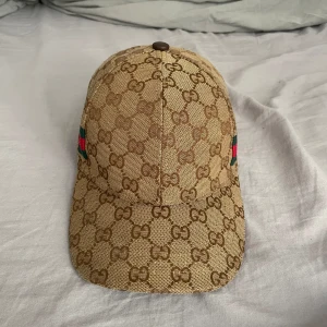 Beige keps gg - Säljer en beige keps med klassiskt GG-monogram över hela kepsen. På sidan finns en grön och röd rand. Kepsen har böjd skärm och justerbar passform. Perfekt för dig som gillar lyxiga accessoarer.